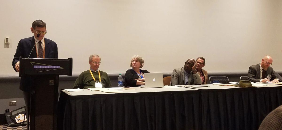 Mark Kinzer speaking at a lectern beside a panel of six seated scholars at a Society for Post-Supersessionist Theology session.